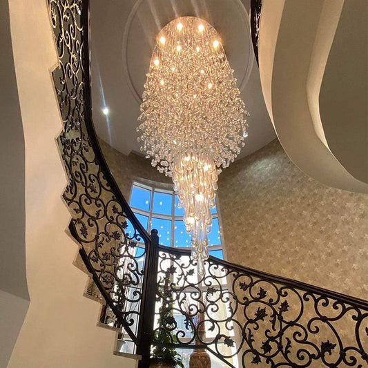 A large, multi-tiered crystal chandelier with warm glowing lights hangs in the center of a grand staircase interior with ornate black metal railings and beige walls.